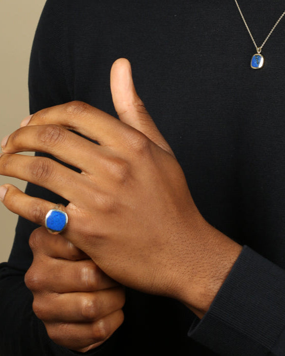 Hand wearing a blue lapis lazuli signet ring with a matching necklace against a black background